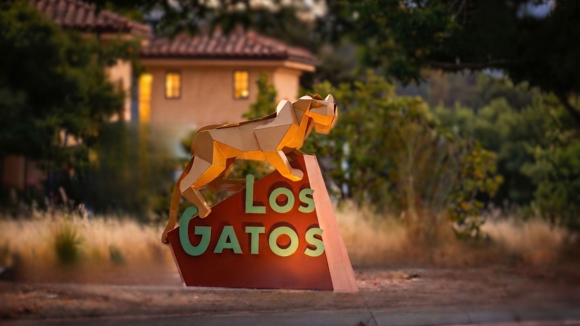 A golden-hour photograph of the Los Gatos sculpture sign surrounded by lush foliage and soft ambient lighting in a luxury real estate setting.