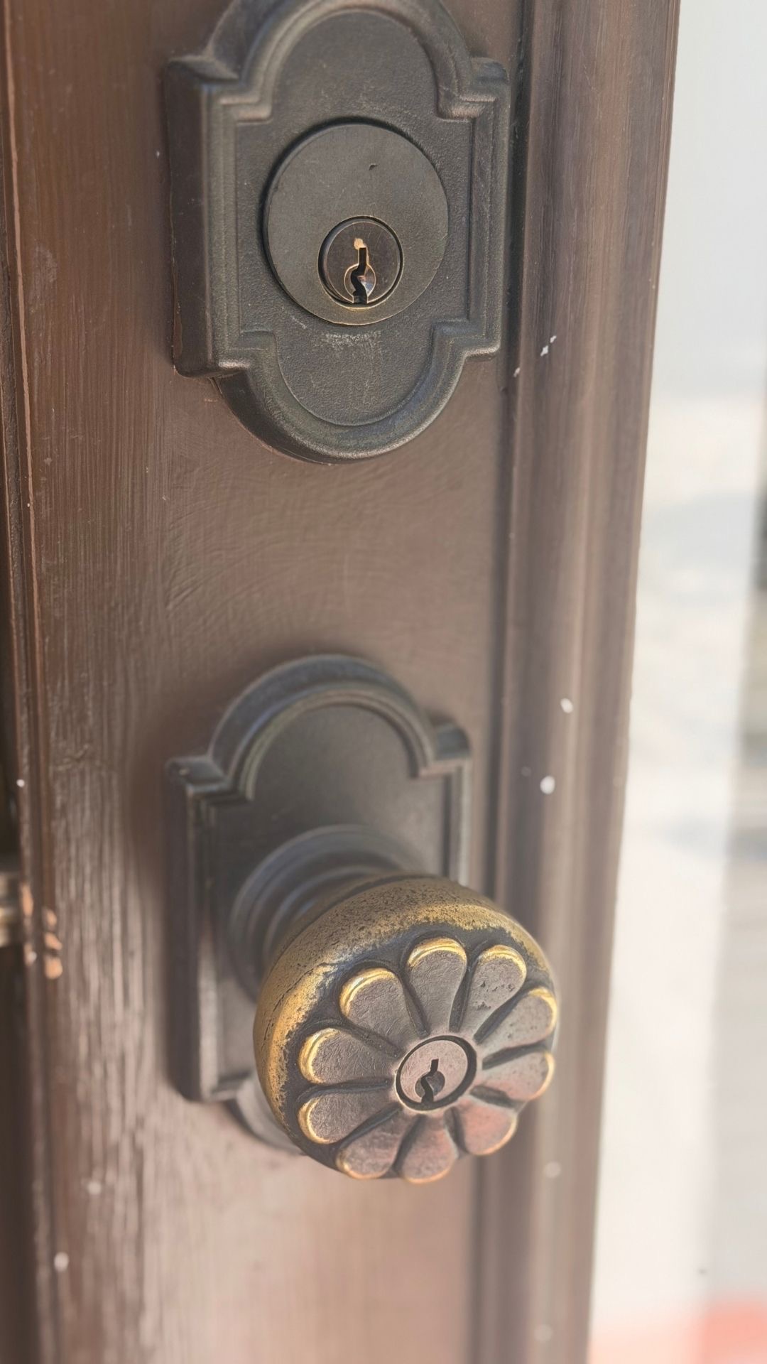 Close-up of an ornate, vintage bronze door knob and deadbolt hardware on a dark solid wood front door.